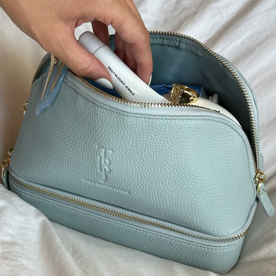Light blue toiletry bag with gold zipper being held open by a hand on a white background